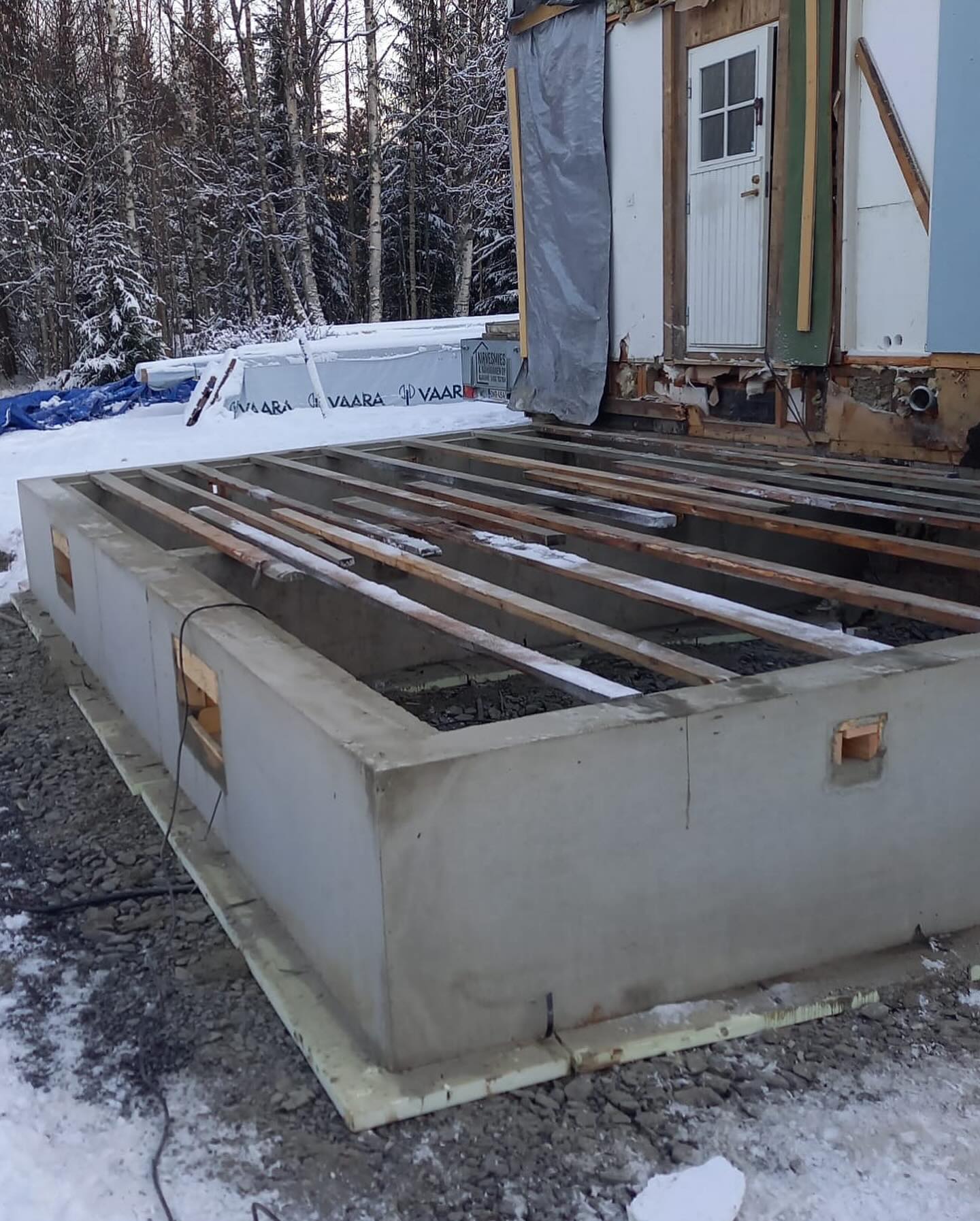 Ulkona on betoninen perustus, jossa on puupalkit ja rakennus, taustalla lumiset puut.