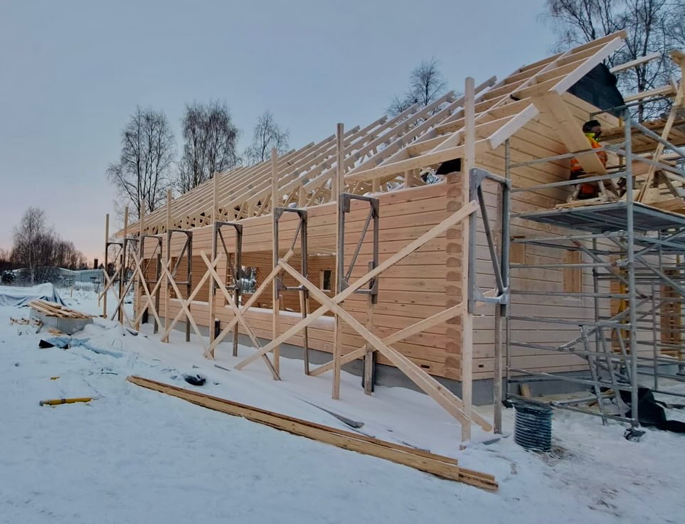 Rakenteilla oleva hirsitalo lumisessa maisemassa, jossa näkyy puurakenteita, telineitä ja työntekijä. Taustalla on puita ja harmaa taivas.