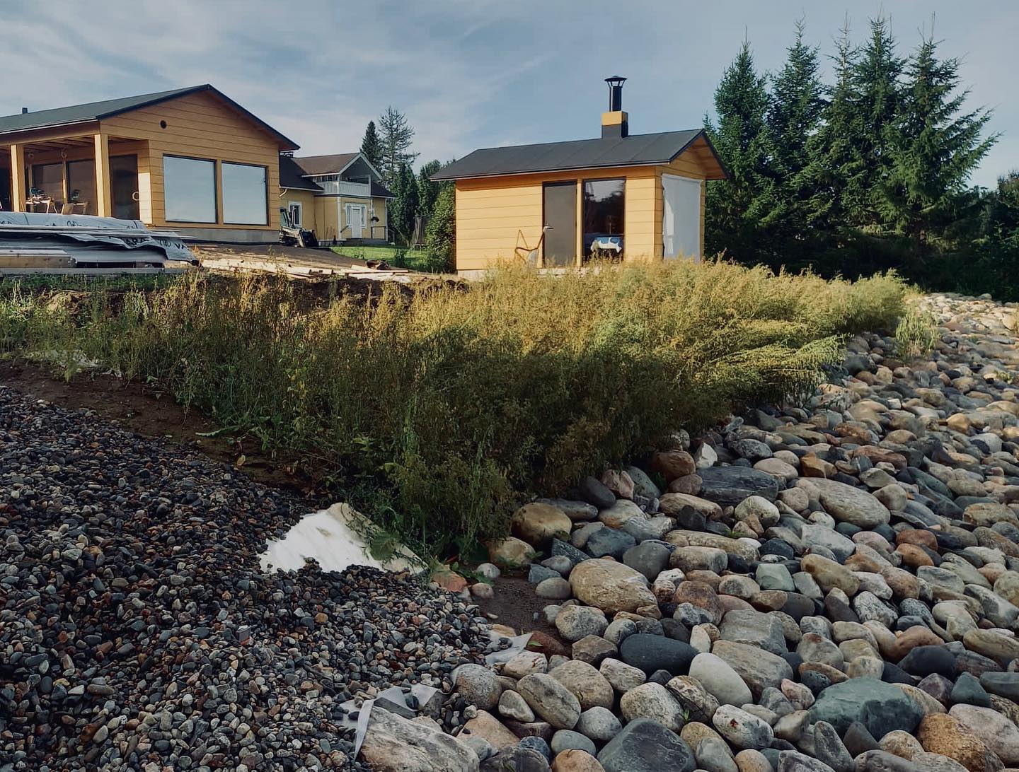 Aurinkoinen maisema, jossa on kaksi keltaista taloa, pieni sauna, kivikkoinen ranta ja vihreitä kasveja.