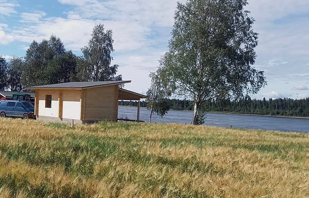 Aurinkoisessa maisemassa on puinen mökki, jonka vieressä kasvaa viljapelto ja taustalla näkyy sininen joki ja metsä.