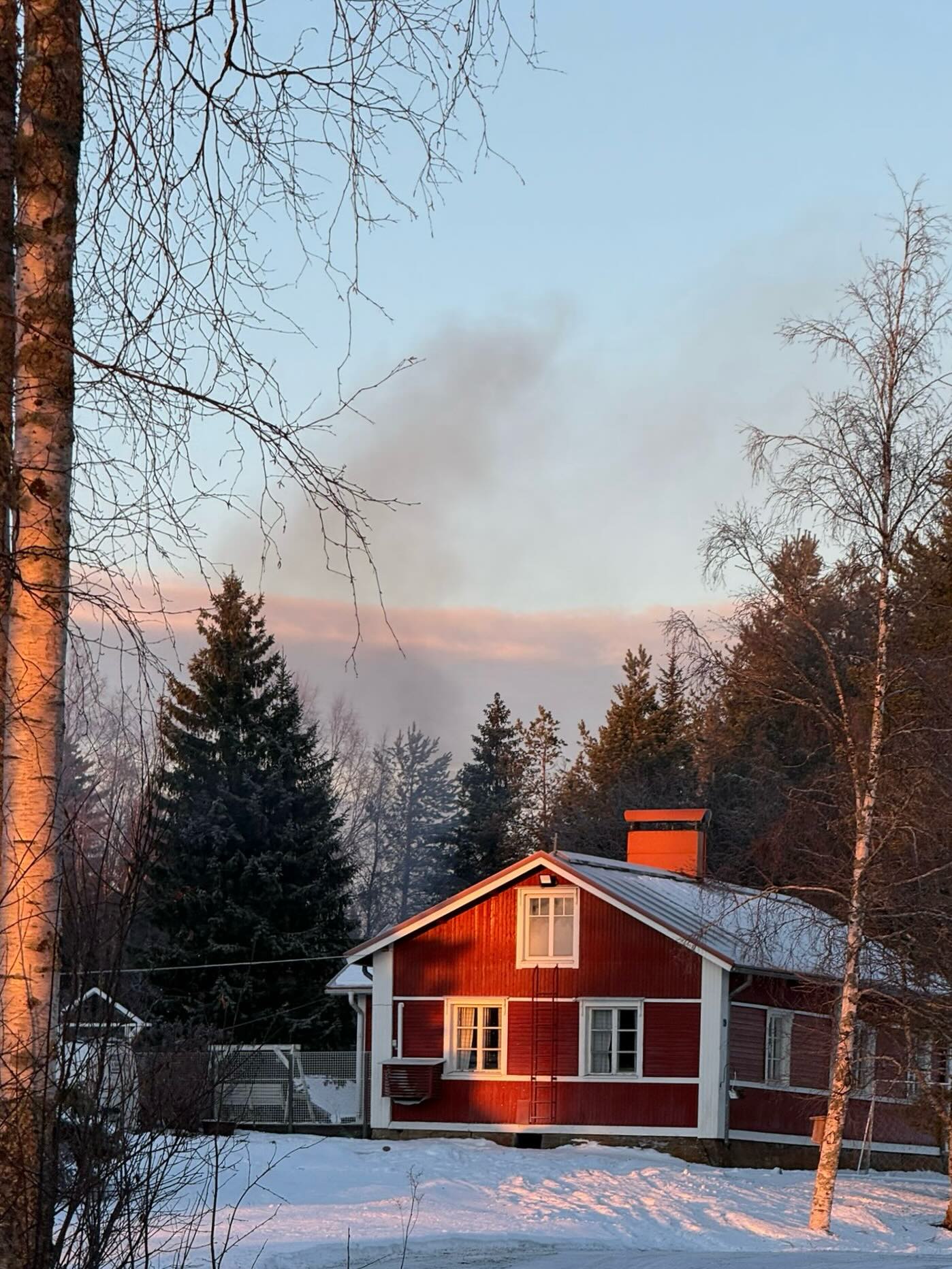 Punainen talo lumisessa maisemassa, auringonlasku valaisee puita ja taivasta. Savua nousee taivaalle.