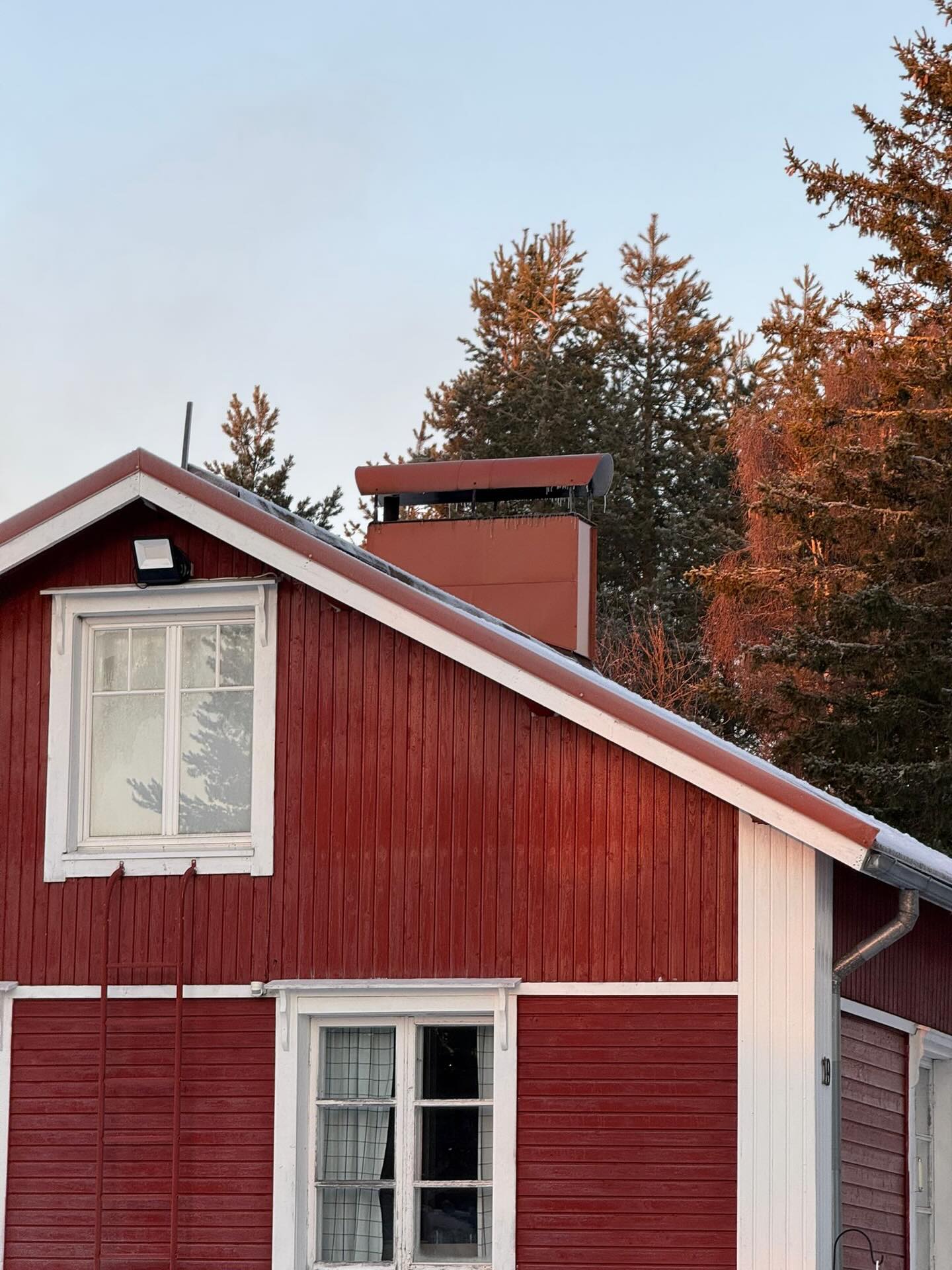 Punainen puutalo, jossa valkoiset ikkunankarmit ja katolla savupiippu, taustalla havupuita ja sininen taivas.