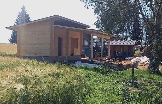 Puurakenteinen rakennus, jossa on katos, sijaitsee pellolla aurinkoisena päivänä. Taustalla näkyy traktori ja toinen rakennus.