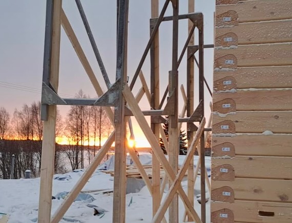 Rakenteilla oleva puutalo lumisessa maisemassa auringonlaskun aikaan, puu- ja metallirakenteet etualalla.