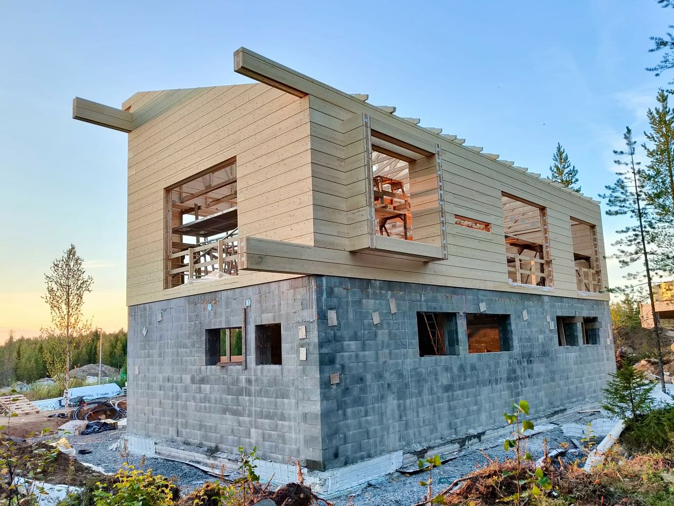 Kaksikerroksinen, rakenteilla oleva puutalo harmaalla kivijalalla, valoisassa ulkoilmassa.