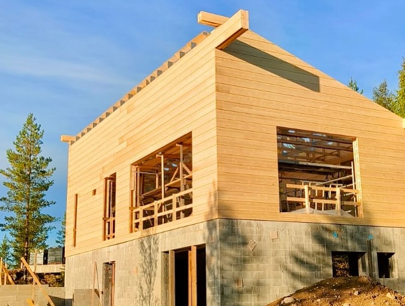 Kaksikerroksinen puutalo rakenteilla, harmaa kivijalka, puuverhous, sininen taivas.