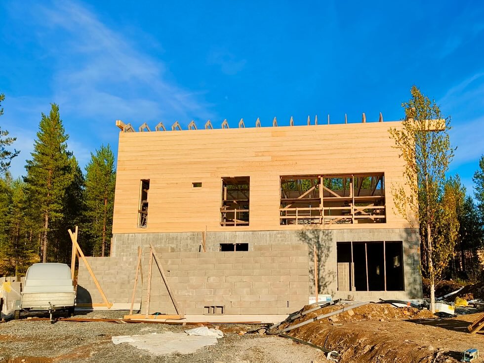 Rakenteilla oleva puutalo, jossa harmaa kivijalka ja puuverhous, metsän ympäröimänä kirkkaassa säässä.