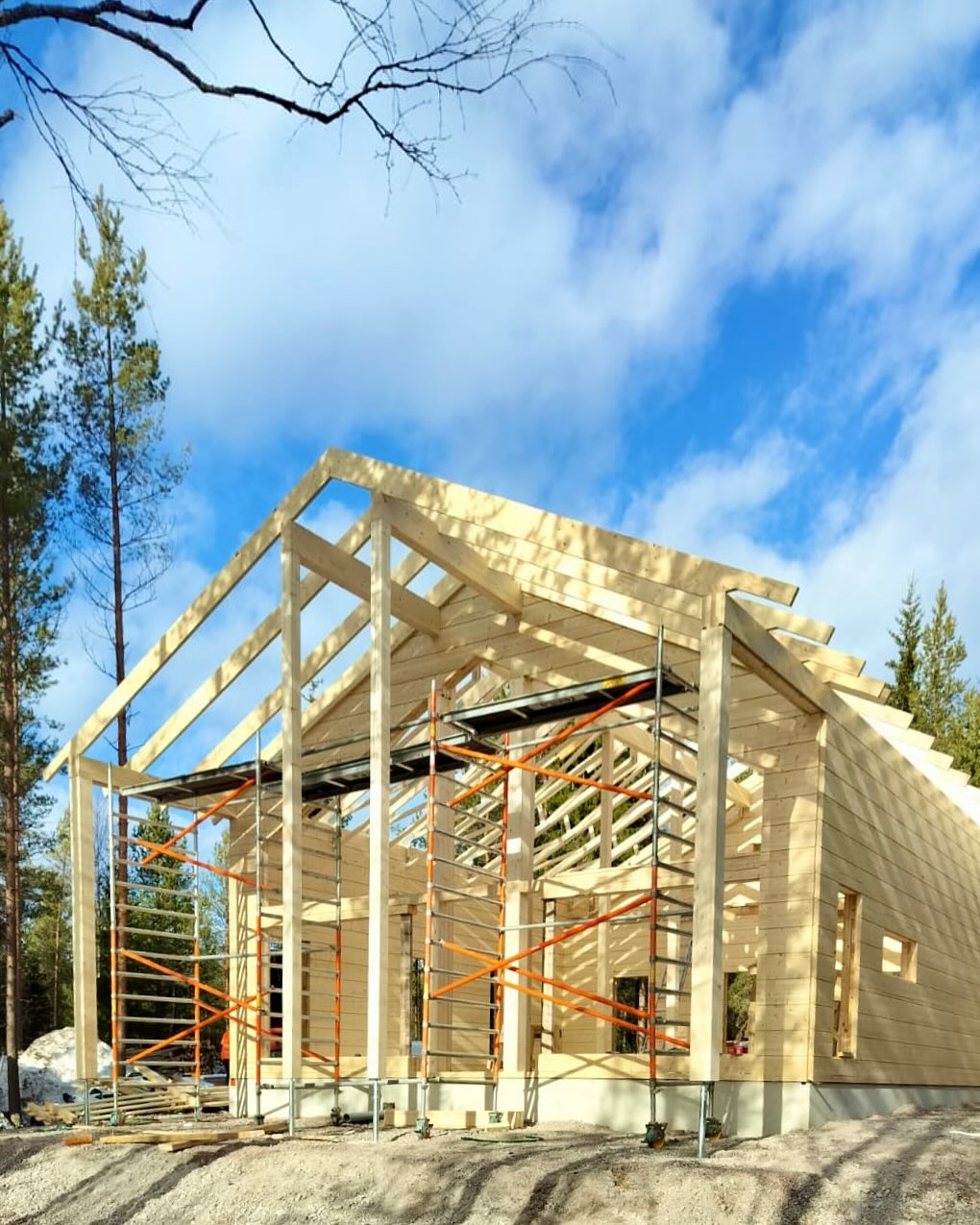 Puurakenteinen talo rakennustelineillä aurinkoisena päivänä, taustalla sininen taivas ja puita.