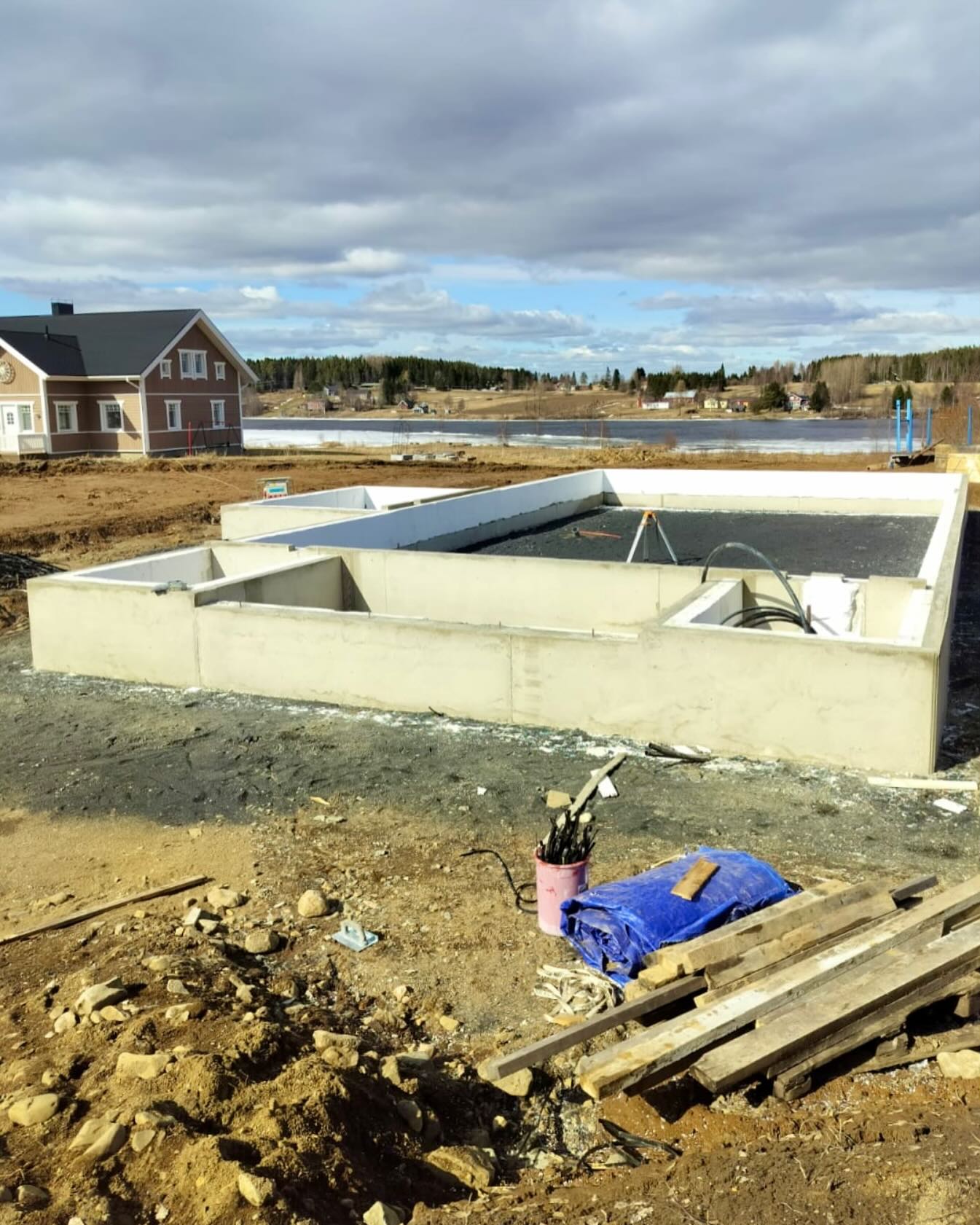 Rakennustyömaa, jossa on valettu betoninen perustus, taustalla talo ja järvimaisema pilvisen taivaan alla.