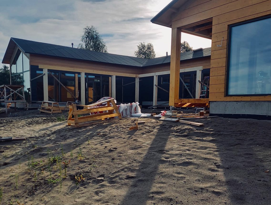 Rakenteilla oleva puutalo, jossa suuret ikkunat ja hiekkainen piha auringonvalossa.