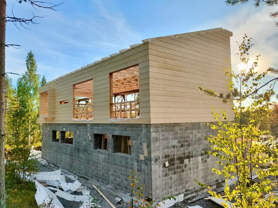 Kaksikerroksinen rakennus, jossa harmaa kivijalka ja puuverhoiltu yläkerta, rakennusvaiheessa metsäisessä ympäristössä aurinkoisena päivänä.