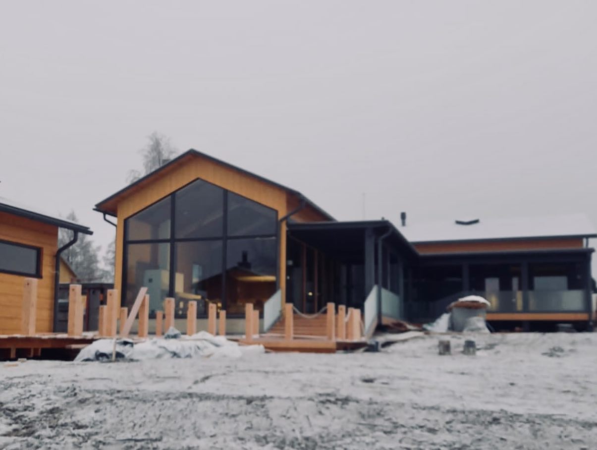 Talvinen maisema esittää puurakenteista taloa, jossa on suuret ikkunat ja lumipeitteinen ympäristö.