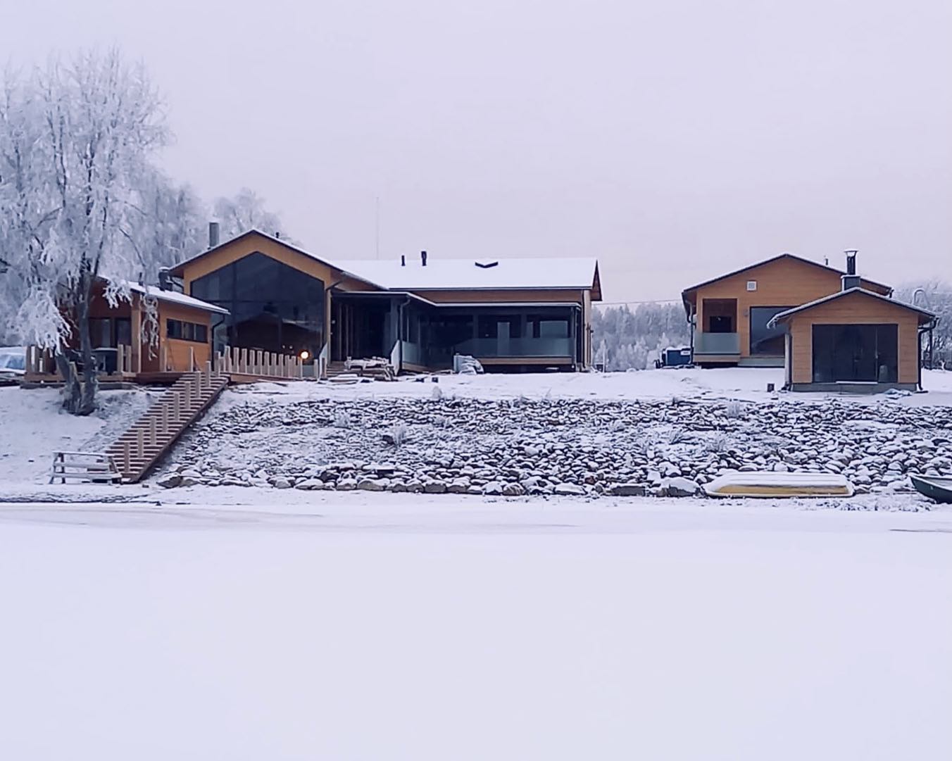 Talvinen maisema, jossa on kolme puista rakennusta lumisella rannalla ja jäätynyt järvi etualalla, harmaa taivas.