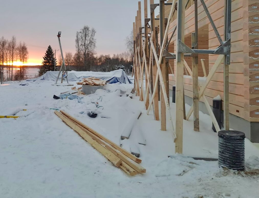 Talvinen rakennustyömaa auringonlaskun aikaan, jossa näkyy puurakenteita, lumipeitteinen maa ja taustalla järvimaisema.
